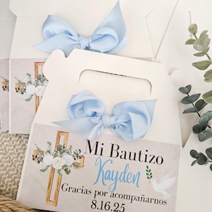May include: A white cardboard favor box with a handle, featuring a light blue satin ribbon bow. The front label displays a wooden cross, white flowers, a dove, and the text Mi Bautizo, Kayden, Gracias por acompañarnos, and 8.16.25.
