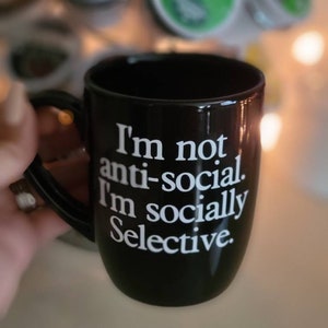 Op de afbeelding: Een zwarte koffiemok met witte tekst die luidt: "Ik ben niet anti-sociaal. Ik ben sociaal selectief."