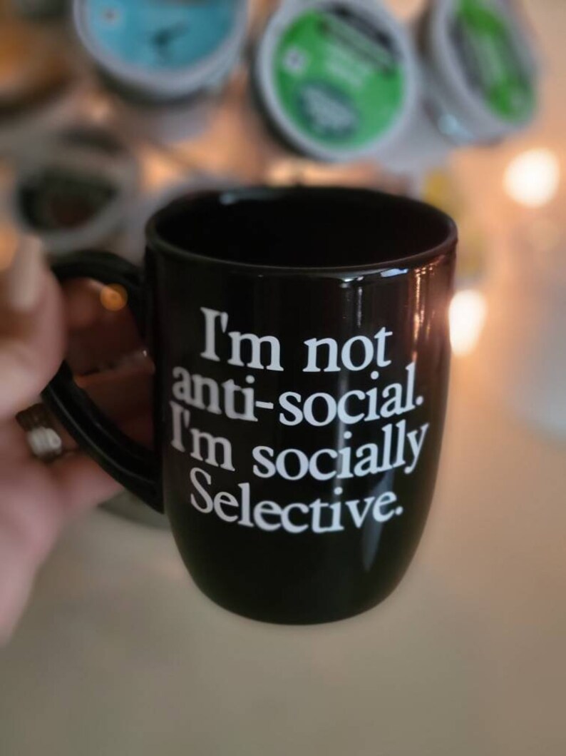 Op de afbeelding: Een zwarte koffiemok met witte tekst die luidt: "Ik ben niet anti-sociaal. Ik ben sociaal selectief."