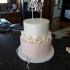 May include: A two-tiered white and pink cake decorated with white roses and a topper that says "God Bless Coral". A silver rosary with pink beads is on the table in front of the cake.