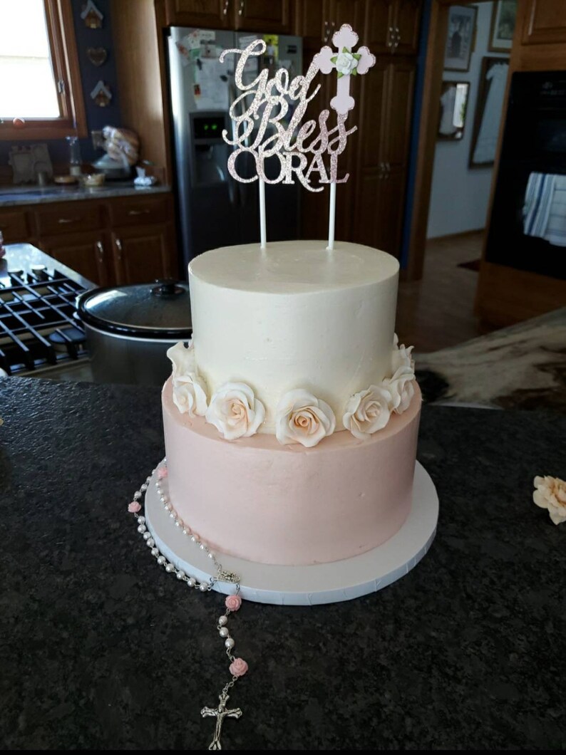 May include: A two-tiered white and pink cake decorated with white roses and a topper that says "God Bless Coral". A silver rosary with pink beads is on the table in front of the cake.