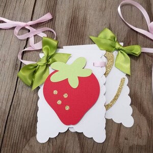 May include: A white banner with a red strawberry and green leaves. The banner is decorated with gold glitter and green bows. The banner is strung with pink ribbon.