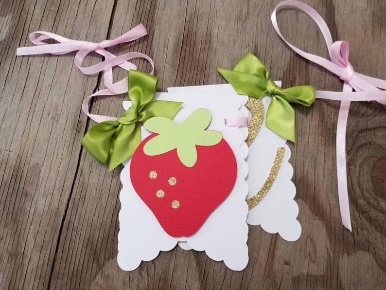 May include: A white banner with a red strawberry and green leaves. The banner is decorated with gold glitter and green bows. The banner is strung with pink ribbon.