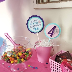 May include: A festive party scene with a pink theme. A clear glass bowl filled with colorful candies sits next to a pink bucket with a decorative sign that says "Happy 4th Birthday Bella". A pink basket holds chocolate candies.