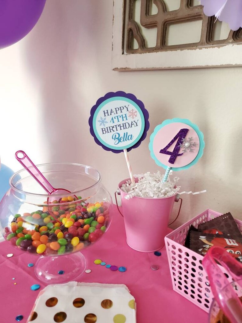 May include: A festive party scene with a pink theme. A clear glass bowl filled with colorful candies sits next to a pink bucket with a decorative sign that says "Happy 4th Birthday Bella". A pink basket holds chocolate candies.