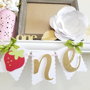 May include: White banner with scalloped edges and green bows. The banner says "one" in gold glitter letters and features a red strawberry with green leaves.