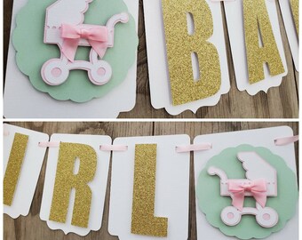 May include: A baby girl banner with the words "Baby Girl" in gold glitter letters. The banner has a green and white background with a pink and white baby carriage design.