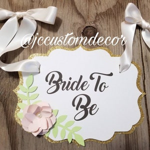May include: White sign with gold glitter trim and a scalloped edge. The sign says "Bride To Be" in black cursive lettering. A pink paper flower with a pearl center and green paper leaves are attached to the sign. Two white satin ribbons are attached to the sign.