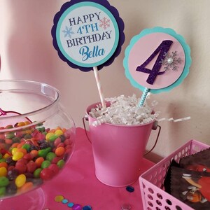 May include: A festive birthday party scene with pink and blue decorations. Two circular signs on sticks read "Happy 4th Birthday Bella" and display the number "4". A pink bucket and a glass bowl filled with colorful candies are on a pink table.