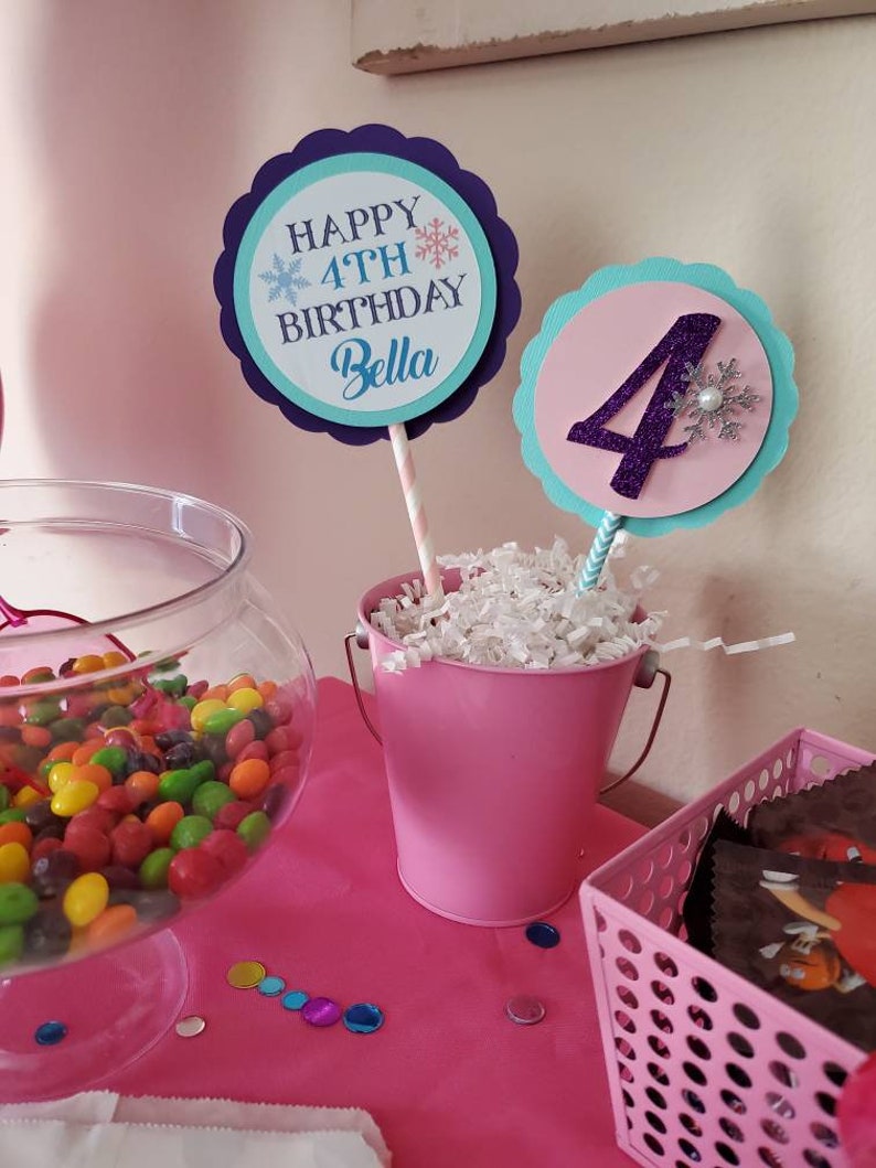 May include: A festive birthday party scene with pink and blue decorations. Two circular signs on sticks read "Happy 4th Birthday Bella" and display the number "4". A pink bucket and a glass bowl filled with colorful candies are on a pink table.