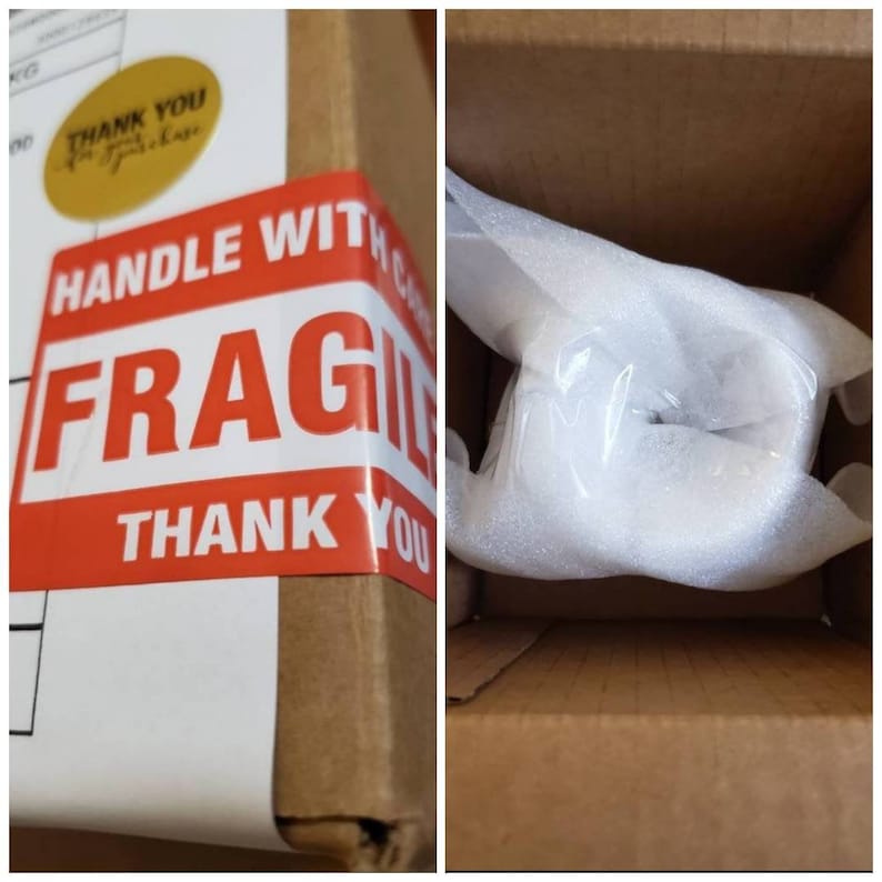 Op de afbeelding: Een bruine kartonnen doos met een rood-witte "HANDLE WITH CARE FRAGILE" sticker en de tekst "THANK YOU" erop gedrukt. De doos is gevuld met witte verpakkingsnoepjes.