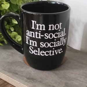 Op de afbeelding: Zwarte keramische koffiemok met witte tekst die "Ik ben niet anti-sociaal. Ik ben sociaal selectief." luidt.