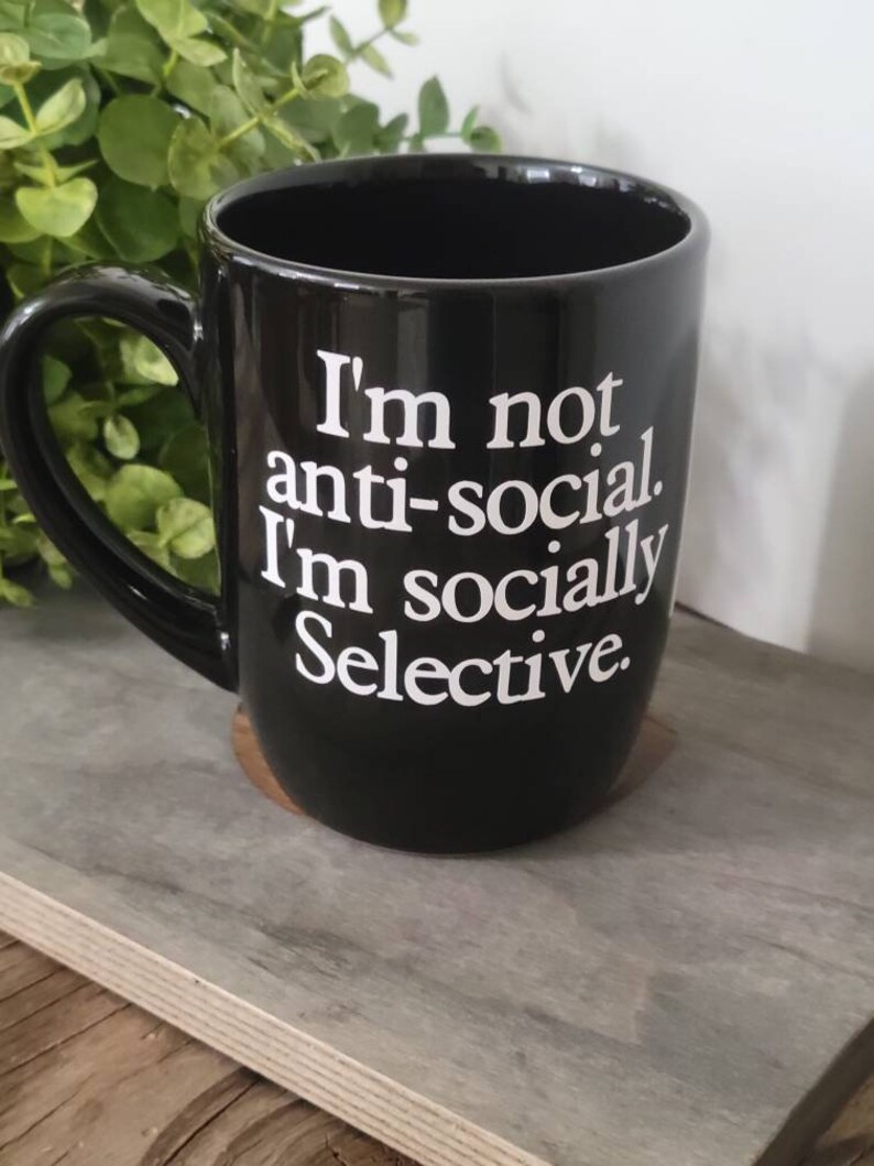 Op de afbeelding: Zwarte keramische koffiemok met witte tekst die "Ik ben niet anti-sociaal. Ik ben sociaal selectief." luidt.