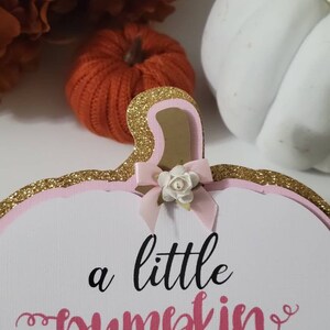Może przedstawiać: Biały znak w kształcie dyni z r&oacute;żowymi i złotymi brokatowymi akcentami. Znak zawiera napis "a little pumpkin is on her way" w czarnym i r&oacute;żowym skrypcie, z małą kokardką i kwiatkiem.