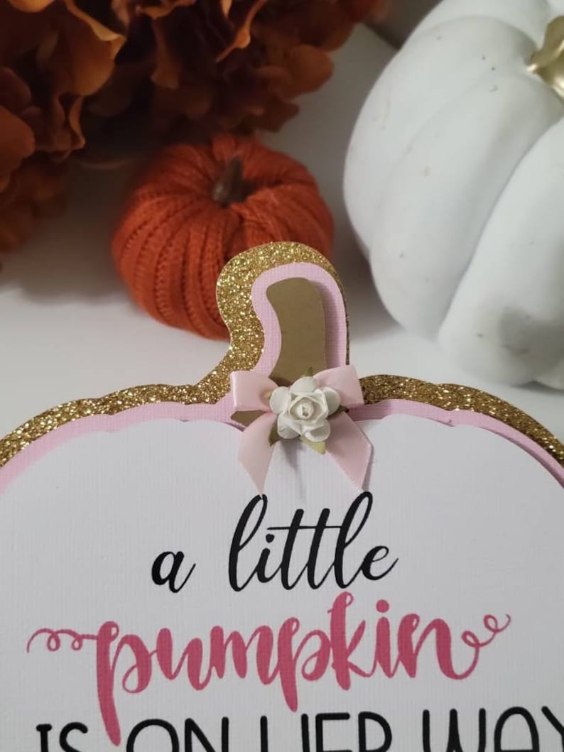 Może przedstawiać: Biały znak w kształcie dyni z r&oacute;żowymi i złotymi brokatowymi akcentami. Znak zawiera napis "a little pumpkin is on her way" w czarnym i r&oacute;żowym skrypcie, z małą kokardką i kwiatkiem.