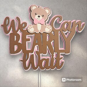 May include: Brown and pink cake topper with the text "We can barely wait" and a brown teddy bear wearing a pink bow tie.