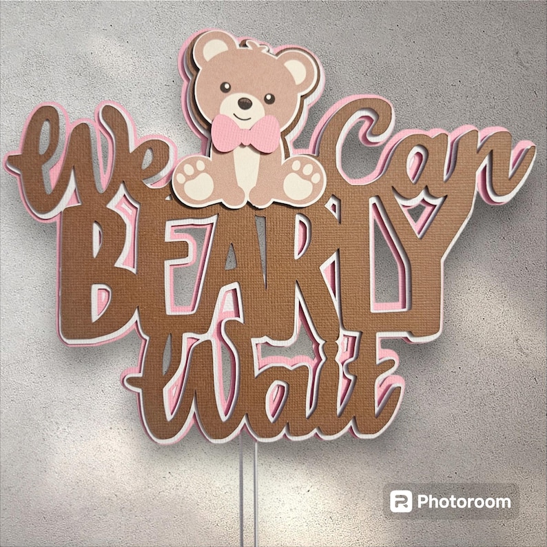 May include: Brown and pink cake topper with the text "We can barely wait" and a brown teddy bear wearing a pink bow tie.