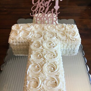 May include: A white cross-shaped cake decorated with white frosting and a pink glitter topper that says "God Bless Piper".