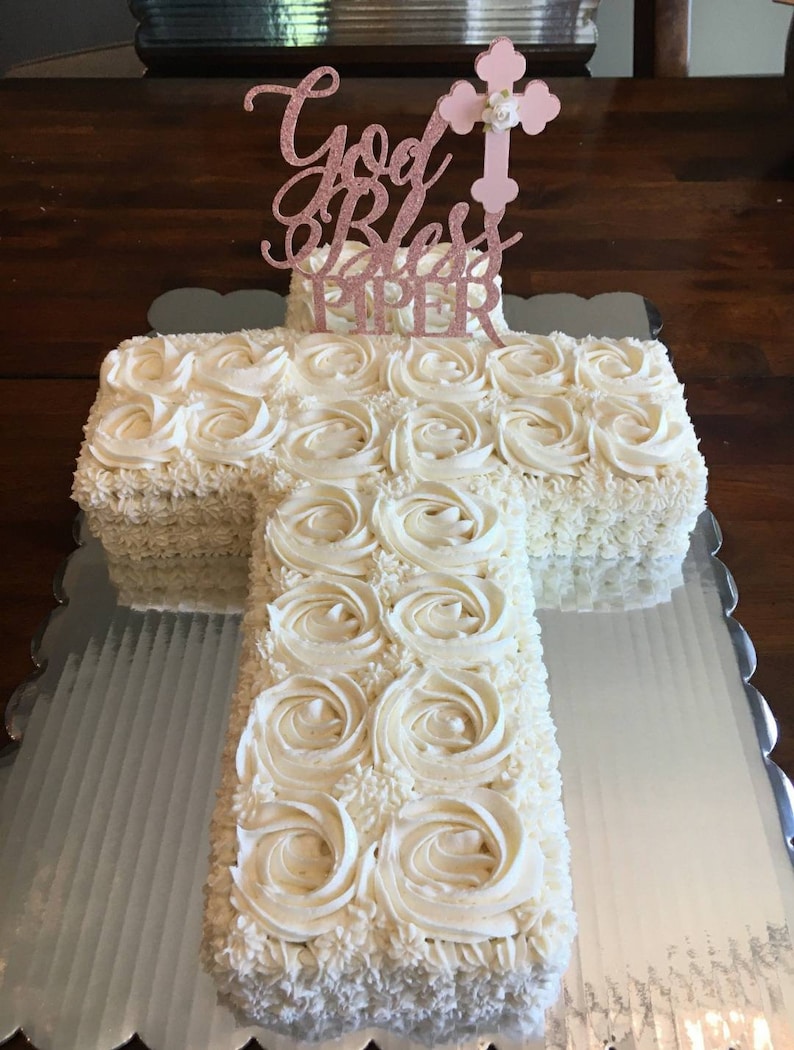 May include: A white cross-shaped cake decorated with white frosting and a pink glitter topper that says "God Bless Piper".