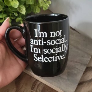 Op de afbeelding: Zwarte keramische koffiemok met witte tekst die "Ik ben niet anti-sociaal. Ik ben sociaal selectief." zegt.