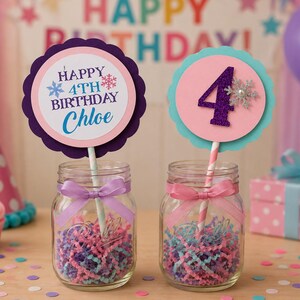 May include: Two glass jars with decorative toppers for a birthday party. One topper says "Happy 4th Birthday Chloe" with snowflakes. The other has a number 4. Both are in jars with colorful paper and ribbons.