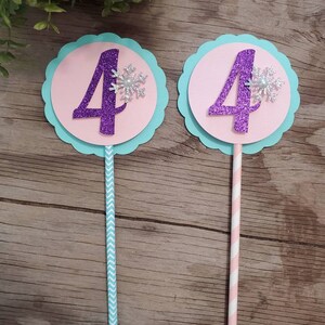 May include: Two birthday cake toppers. Each has a purple glitter number 4 on a pink circle, a silver snowflake, and a light blue scalloped edge. One has a blue chevron straw, the other a pink and white striped straw.