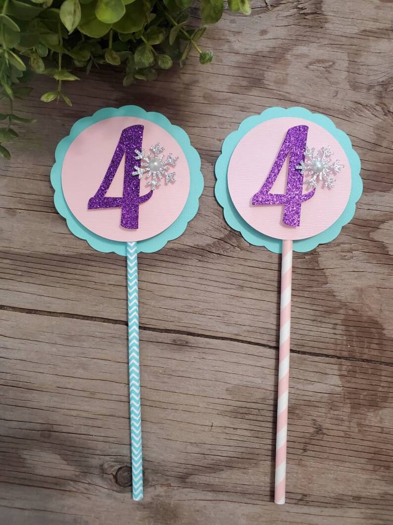 May include: Two birthday cake toppers. Each has a purple glitter number 4 on a pink circle, a silver snowflake, and a light blue scalloped edge. One has a blue chevron straw, the other a pink and white striped straw.