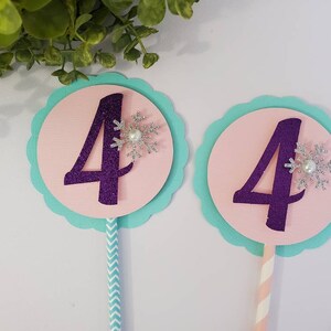 May include: Two birthday cake toppers with the number 4 in purple glitter. Each topper has a pink circle, a light blue scalloped edge, and a silver snowflake with a pearl. One has a blue and white striped stick, the other a pink and white striped stick.