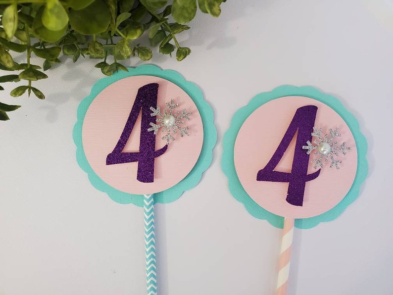 May include: Two birthday cake toppers with the number 4 in purple glitter. Each topper has a pink circle, a light blue scalloped edge, and a silver snowflake with a pearl. One has a blue and white striped stick, the other a pink and white striped stick.
