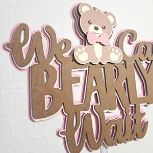 May include: A brown and pink layered cake topper with the text "We Can't Wait" and a brown teddy bear wearing a pink bow tie.
