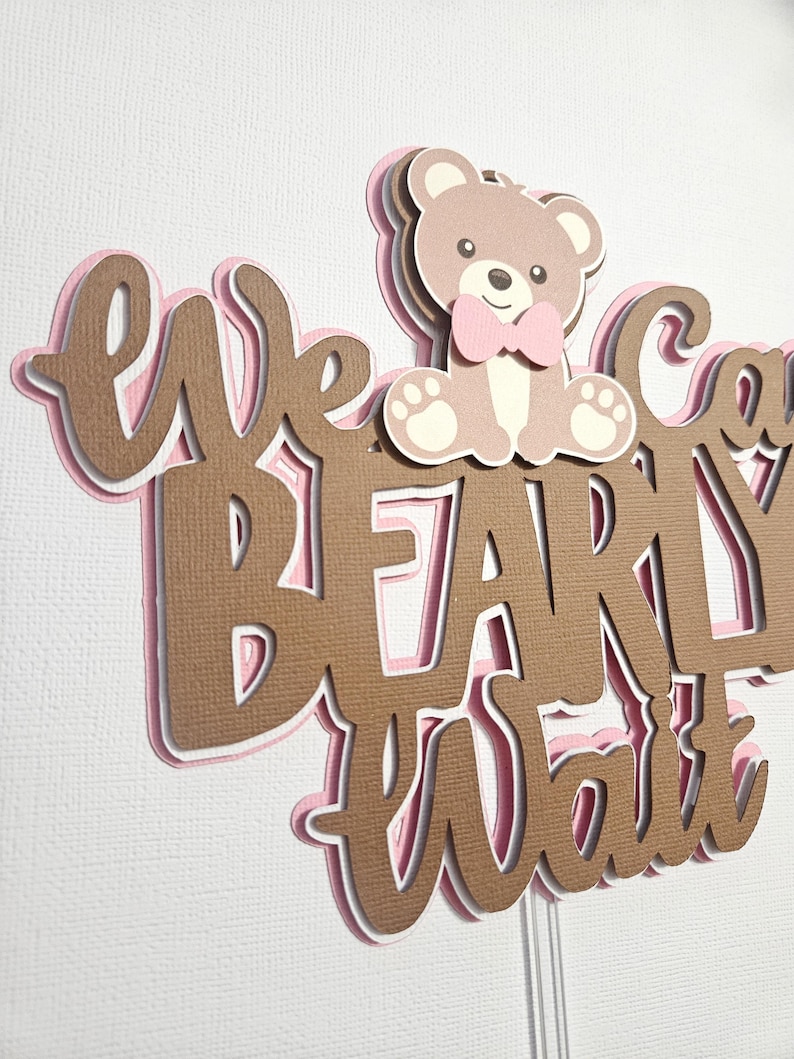 May include: A brown and pink layered cake topper with the text "We Can't Wait" and a brown teddy bear wearing a pink bow tie.