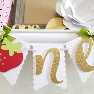 May include: A white banner with scalloped edges and gold glitter letters spelling "one". The banner is decorated with a red strawberry, green leaves, and green bows.