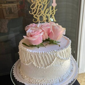 May include: White cake with white frosting decorated with a gold glitter topper that says "God Bless Sofia" and a pink cross with a white flower. The cake is topped with pink roses.
