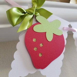 May include: A red and green paper strawberry with gold glitter accents, attached to a white scalloped paper banner with a green satin bow.
