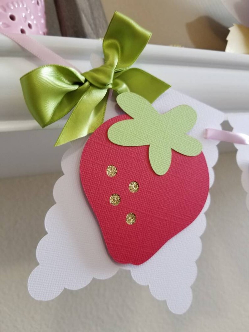 May include: A red and green paper strawberry with gold glitter accents, attached to a white scalloped paper banner with a green satin bow.