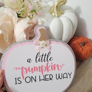 Może przedstawiać: Ozdobny znak w kształcie dyni z napisem "a little pumpkin is on her way" w kolorze r&oacute;żowym i czarnym. Znak ma r&oacute;żową obw&oacute;dkę ze srebrnym brokatem i małą kokardką. Biała dynia ze złotą łodygą i pomarańczowa dynia w tle.