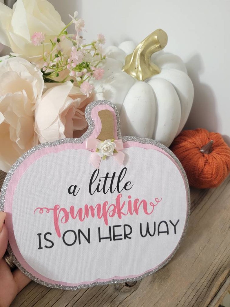 Może przedstawiać: Ozdobny znak w kształcie dyni z napisem "a little pumpkin is on her way" w kolorze r&oacute;żowym i czarnym. Znak ma r&oacute;żową obw&oacute;dkę ze srebrnym brokatem i małą kokardką. Biała dynia ze złotą łodygą i pomarańczowa dynia w tle.