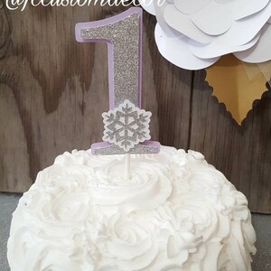 May include: A white cake with a silver glitter snowflake and a purple number one topper. The cake is decorated with white frosting and has a white flower in the background.
