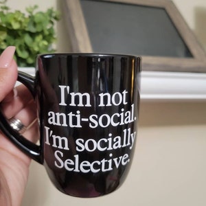 Op de afbeelding: Een zwarte koffiemok met witte tekst die luidt: "Ik ben niet anti-sociaal. Ik ben sociaal selectief."