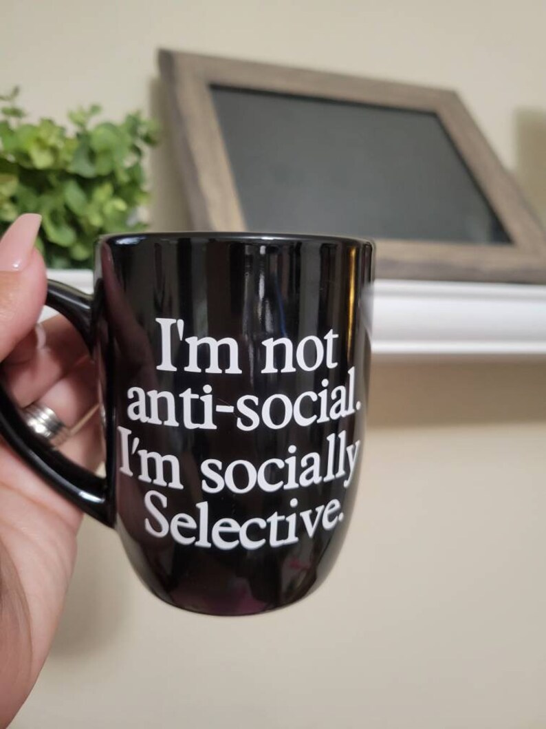 Op de afbeelding: Een zwarte koffiemok met witte tekst die luidt: "Ik ben niet anti-sociaal. Ik ben sociaal selectief."