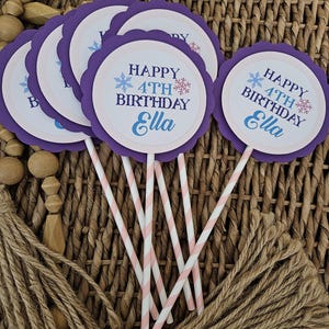 May include: A collection of purple and white cupcake toppers with the text "HAPPY 4TH BIRTHDAY Ella" in blue and white. The toppers have a scalloped edge and are attached to pink and white striped paper straws. The background is a woven basket.