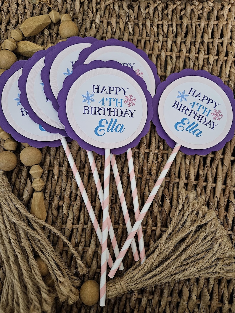 May include: A collection of purple and white cupcake toppers with the text "HAPPY 4TH BIRTHDAY Ella" in blue and white. The toppers have a scalloped edge and are attached to pink and white striped paper straws. The background is a woven basket.