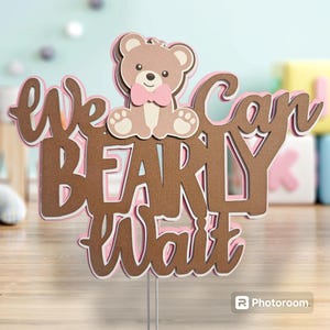 May include: A brown and pink cake topper with the text "We can barely wait" and a brown teddy bear wearing a pink bow tie.