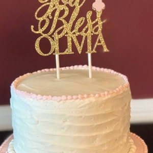 May include: A two-tiered cake decorated with pink frosting and white pearl accents. A gold glitter cake topper with the text "God Bless Olivia" and a pink cross with a white flower is on top of the cake.