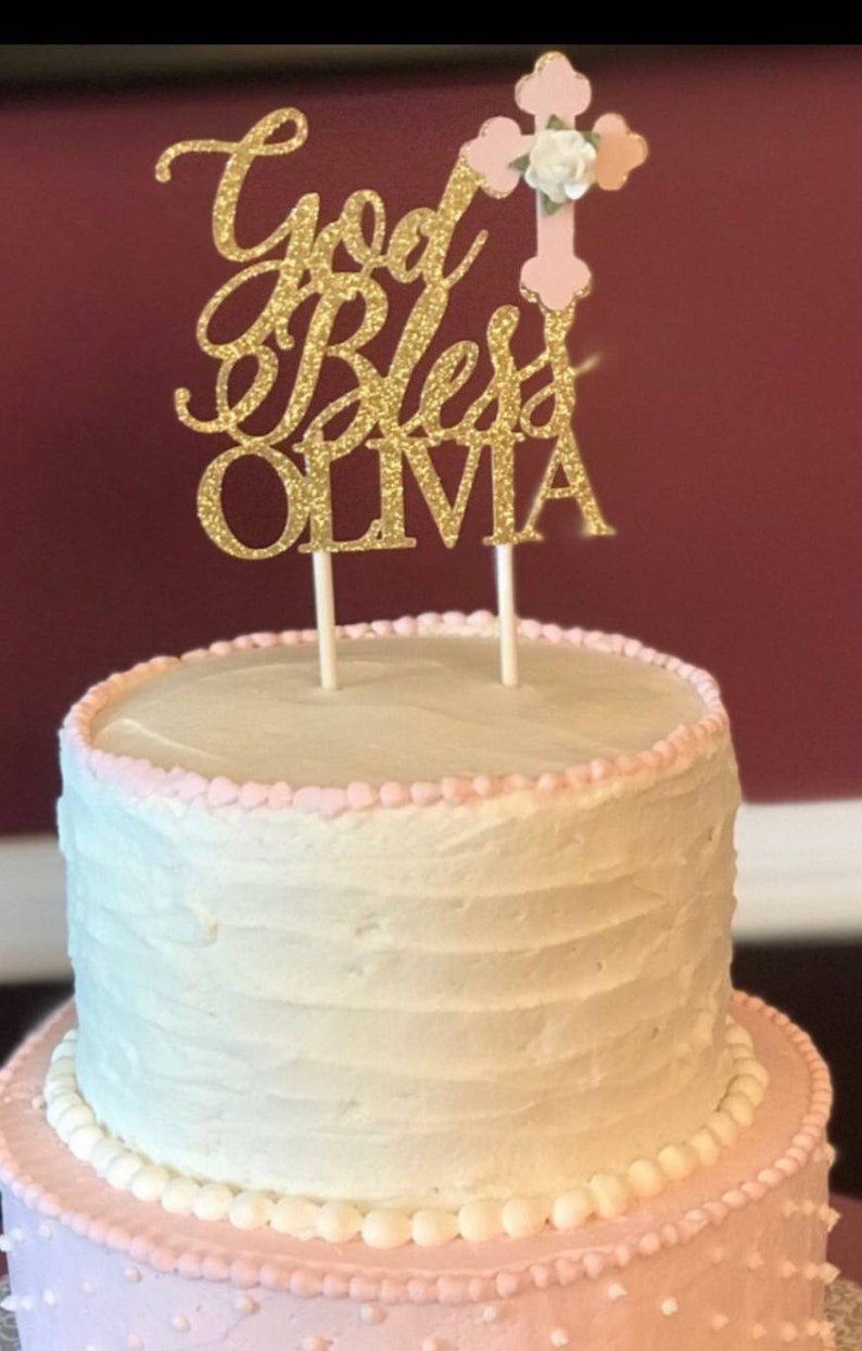May include: A two-tiered cake decorated with pink frosting and white pearl accents. A gold glitter cake topper with the text "God Bless Olivia" and a pink cross with a white flower is on top of the cake.