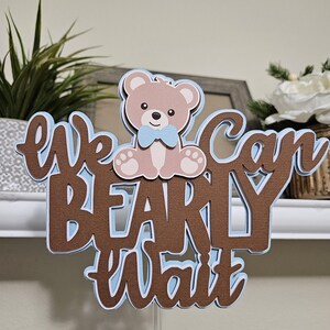 May include: Brown and blue cake topper with the text "We can barely wait" and a teddy bear wearing a blue bow tie.