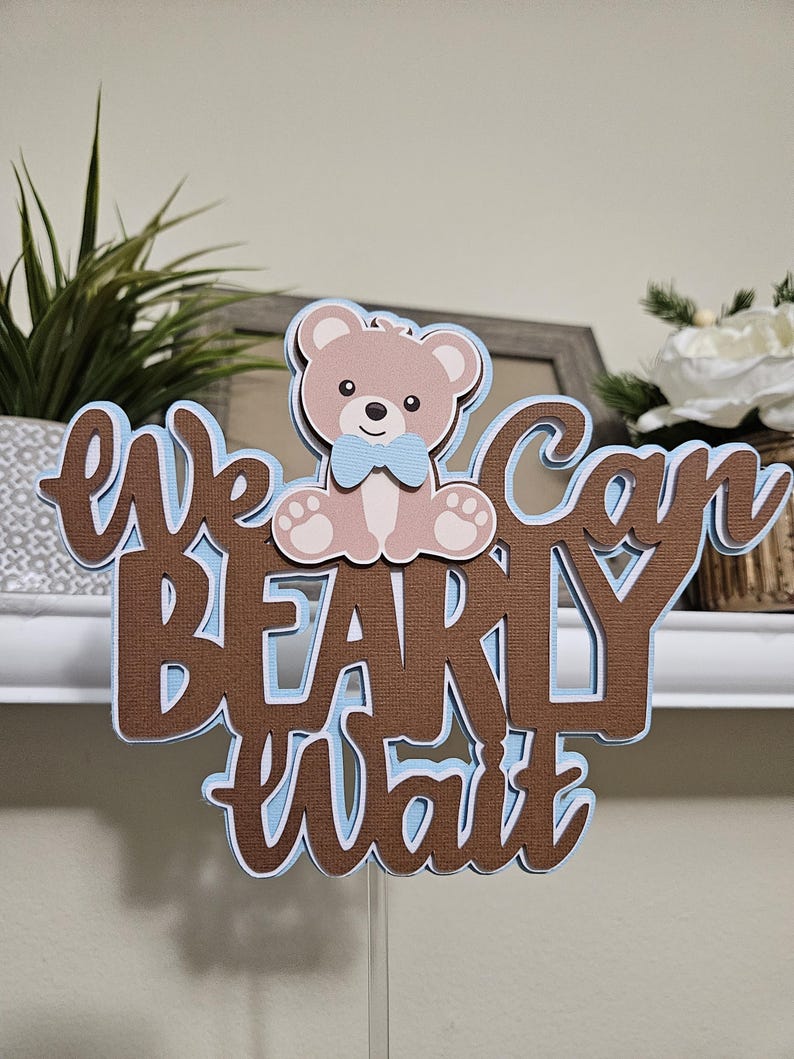 May include: Brown and blue cake topper with the text "We can barely wait" and a teddy bear wearing a blue bow tie.