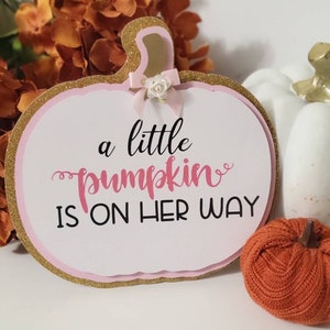 Może przedstawiać: Ozdobny znak w kształcie dyni z napisem "a little pumpkin is on her way". Znak ma złotą, błyszczącą obw&oacute;dkę, r&oacute;żowe akcenty i małą kokardkę. Widoczna jest r&oacute;wnież pomarańczowa i biała dynia.