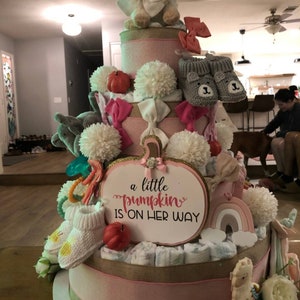 Może przedstawiać: Wielopoziomowy tort z pieluch na baby shower, ozdobiony r&oacute;żowymi wstążkami, kokardkami i pluszowymi zwierzętami. Tort zawiera napis "a little pumpkin is on her way". Zawiera buciki dla dziecka, pompony i małe dynie.
