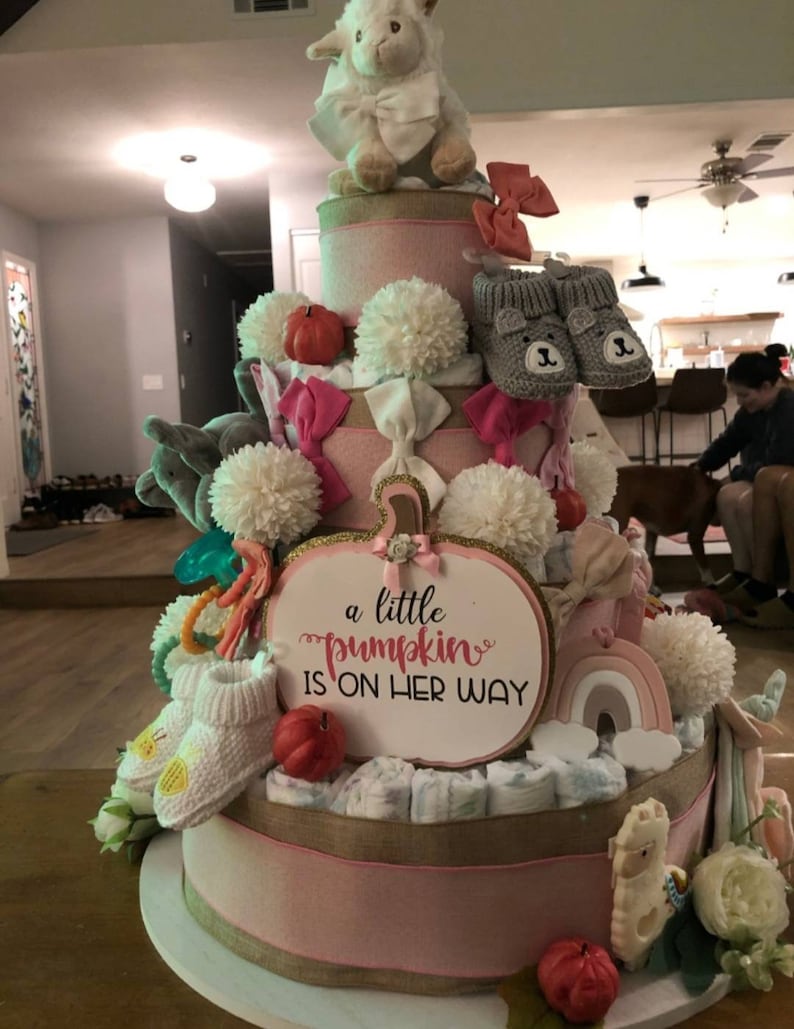 Może przedstawiać: Wielopoziomowy tort z pieluch na baby shower, ozdobiony r&oacute;żowymi wstążkami, kokardkami i pluszowymi zwierzętami. Tort zawiera napis "a little pumpkin is on her way". Zawiera buciki dla dziecka, pompony i małe dynie.
